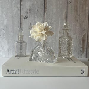 May include: Three clear glass bottles of varying shapes and textures, with one triangular bottle featuring a large, cream-coloured artificial flower. The bottles are arranged on a white book with the text Artful Lifestyle printed on the spine.