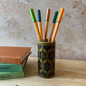 May include: A cylindrical ceramic pen holder with a dark green and brown geometric pattern containing five coloured felt-tip pens in shades of lime green, teal, tan, dark green, and orange, positioned next to two stacked books with orange and green covers.
