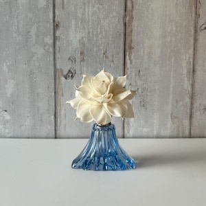 May include: A translucent blue glass bottle with a fluted, flared base and a silver-toned collar. A cream-coloured, multi-layered flower made of thin, delicate petals is positioned at the top of the bottle, serving as a decorative topper.