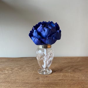 May include: A striking blue flower displayed in a small, clear crystal vase. The vase features a silver rim and a diamond-cut design. The flower's intense colour provides a strong contrast against the clear glass.