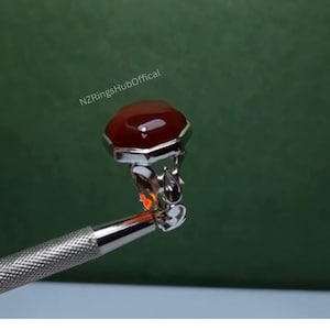 May include: A silver ring with a large, octagonal, reddish-brown gemstone. The ring is held by a silver tool against a green background. The text "NZRingsHubOffical" is visible in the upper left corner.