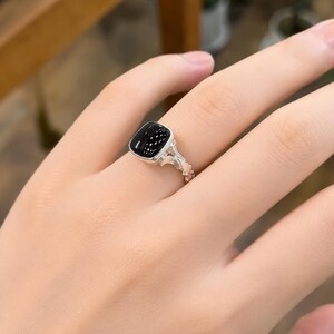 May include: A silver ring featuring a square, black gemstone with a speckled appearance. The ring's band has an ornate, textured design. The ring is shown on a person's finger.