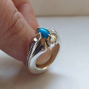 Op de afbeelding: Een zilveren ring met een heldere turquoise ovale steen. De ring heeft een gedetailleerd ontwerp met een textuurpatroon aan de zijkanten. De ring wordt op een vinger getoond, wat het vakmanschap en de kleur van het sieraad benadrukt.