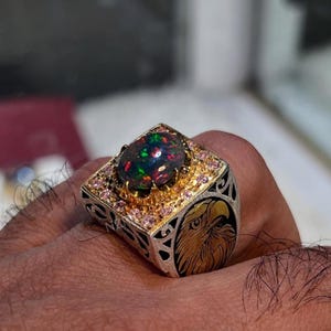 May include: A silver ring with a large, oval black opal gemstone. The ring has gold accents, small pink gemstones, and an engraved eagle design on the side. The opal displays a vibrant play of colour.