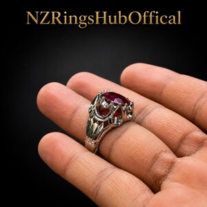 Könnte beinhalten: Ein silberfarbener Ring mit einem großen, tiefroten facettierten Edelstein, der in einer komplexen, durchbrochenen Metallfassung sitzt. Der Text NZRingsHubOffical steht in goldener Schrift über dem Ring vor schwarzem Hintergrund.