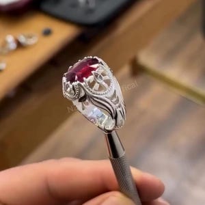 Könnte beinhalten: Ein silberner Ring mit einem ovalen, tiefroten Edelstein. Der Ring hat ein aufwendiges, detailliertes Design um den Stein und am Band. Der Ring wird von einem Metallwerkzeug gehalten, möglicherweise zur Präsentation.