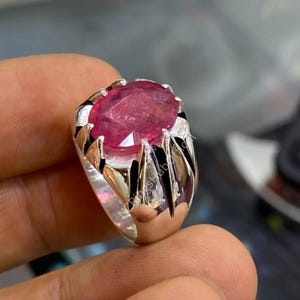Könnte beinhalten: Ein silberner Ring mit einem großen, oval geschliffenen Rubin. Der Rubin hat eine tiefe, lebendige rote Farbe und ist in einem polierten Silberband mit dekorativen Akzenten gefasst. Der Ring hat ein kühnes, maskulines Design.