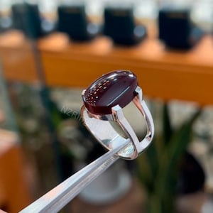 Könnte beinhalten: Ein silberner Ring mit einem großen, ovalen, dunkelroten Edelstein. Der Edelstein hat ein gesprenkeltes Muster. Der Ring wird von einer Pinzette gehalten. Der Markenname "NZRingsHub" ist sichtbar.