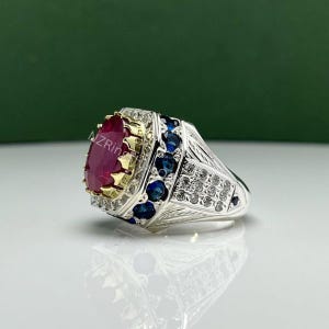 Könnte beinhalten: Ein silberner Ring mit einem großen, ovalen, roten Edelstein als Mittelpunkt. Der Ring ist mit Gold, blauen und klaren Steinen verziert. Der Text "AZRing" ist auf dem Edelstein sichtbar.