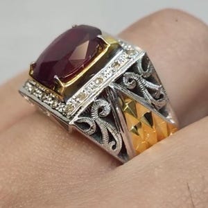 May include: A close-up of a silver and gold ring featuring a large, rectangular, deep red gemstone. The ring has intricate filigree detailing and small accent stones around the gemstone setting.