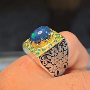 May include: A silver ring with a large oval, dark blue opal gemstone. The ring features a gold setting with small green stones and a floral engraved design on the sides. The text "NZRingShop" is visible on the ring.