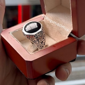 May include: A silver ring featuring a large, oval black stone, set within a floral pattern. The ring is presented in a red wooden box with a cream-coloured lining. The text "NZRingsHubOffical" is visible.