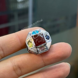 May include: A silver ring with a central rectangular red stone, inscribed with text. The ring is surrounded by smaller gemstones in various colors, including blue, green, and pink. The ring has an octagonal design.