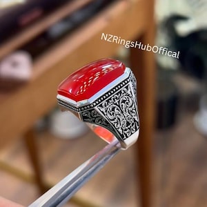 May include: A silver ring featuring a red gemstone and detailed floral engravings. The ring is set on a silver band and has a square-shaped gemstone.