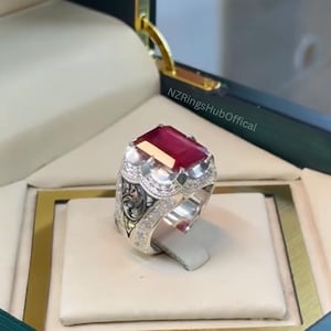 Puede incluir: Un anillo de plata con una gran gema rectangular de color rojo intenso. El anillo tiene detalles intrincados alrededor de la gema y a lo largo de la banda, con un patrón decorativo en los lados. El anillo se muestra en una caja de joyería.