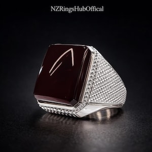 May include: A silver ring with a large, rectangular, dark red stone. The stone has a white design. The ring's band is encrusted with small, clear stones. The text "NZRingsHubOffical" is at the top.