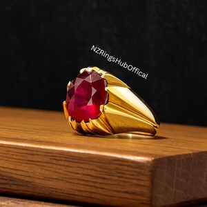 May include: A gold ring featuring a large, oval-cut, deep red gemstone. The ring is displayed on a wooden surface. The text "NZRingsHubOffical" is visible in the background.