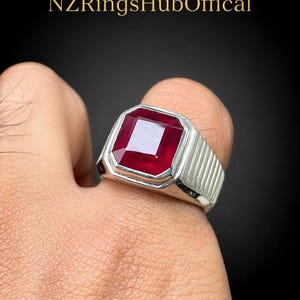 May include: A silver ring with a large, rectangular, deep red gemstone. The ring has a ribbed design on the band and is set against a dark background. The text "NZRingsHubOffical" is at the top.