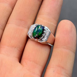 May include: A silver ring with an oval, black opal gemstone with green and blue iridescence. The ring is set with small, clear accent stones and has a rectangular design. The text "NZRingsHubOfficial" is visible.