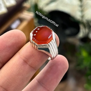 May include: A silver ring with a large, octagonal, reddish-orange gemstone. The ring has a detailed, ribbed design on the sides and is held by a person's fingers. The text "NZRingsHubOffical" is visible.