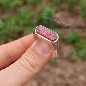 May include: A silver ring with a rectangular, red stone. The stone features Arabic calligraphy. The ring is held in a hand against a blurred green background. The text "NZRingsHubOffical" is visible.