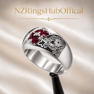 Puede incluir: Un anillo de tono plateado con un grupo de cuatro piedras de corte ovalado en rojo intenso engastadas en una sección elevada. La banda muestra grabados intrincados de flores y volutas rellenos de negro. El texto NZRingsHubOffical aparece arriba.