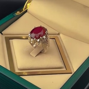 Peut inclure: Une bague en argent ornée d'une pierre précieuse ovale rouge foncé. La bague présente un design orné et est présentée dans un écrin vert et beige.