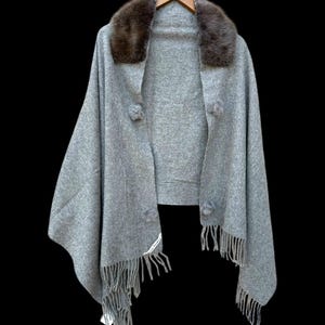 Op de afbeelding: Een grijze cape met een bruine bontkraag, gepresenteerd op een houten hanger tegen een zwarte achtergrond. De cape heeft pompons en franjes. Een label met witte tekst is zichtbaar. Een stijlvol kledingstuk.