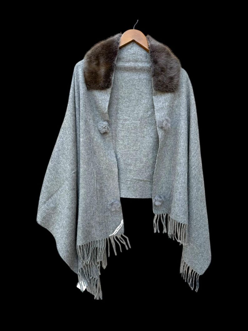 Op de afbeelding: Een grijze cape met een bruine bontkraag, gepresenteerd op een houten hanger tegen een zwarte achtergrond. De cape heeft pompons en franjes. Een label met witte tekst is zichtbaar. Een stijlvol kledingstuk.