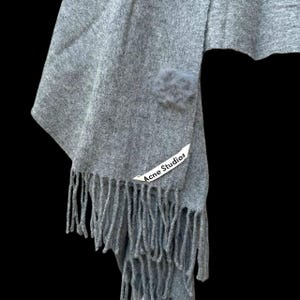Op de afbeelding: Grijze sjaal met franjes en een kleine, pluizige pompon. De sjaal heeft een label met de tekst "Acne Studios". De sjaal is gemaakt van een zacht materiaal.