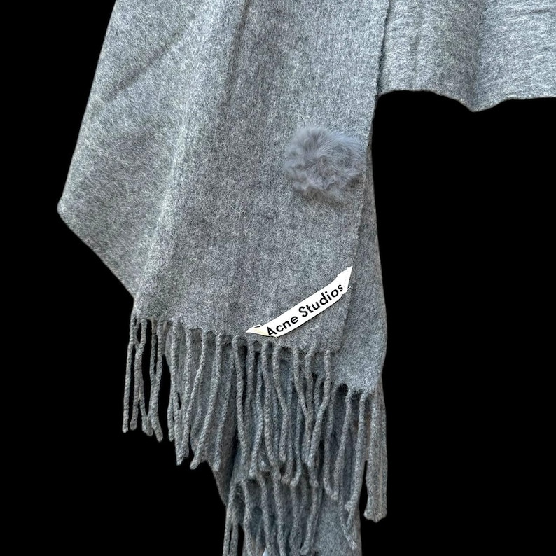 Op de afbeelding: Grijze sjaal met franjes en een kleine, pluizige pompon. De sjaal heeft een label met de tekst "Acne Studios". De sjaal is gemaakt van een zacht materiaal.