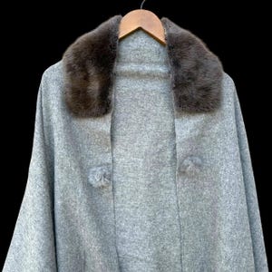 Op de afbeelding: Een grijze cape met een donkerbruine imitatiebontkraag en twee bijpassende pomponsluitingen. De cape hangt aan een houten kleerhanger tegen een zwarte achtergrond. De stof lijkt een zacht, wolachtig materiaal te zijn.