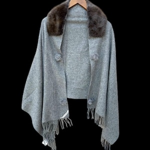 Op de afbeelding: Een grijze cape met een bruine bontkraag, hangend aan een houten hanger tegen een zwarte achtergrond. De cape heeft drie decoratieve pompons en franjes langs de onderkant. Een label is zichtbaar op de franje.