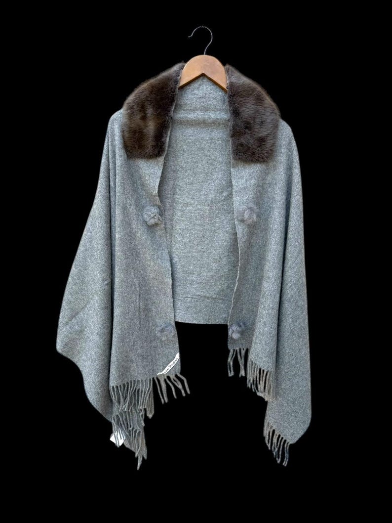 Op de afbeelding: Een grijze cape met een bruine bontkraag, hangend aan een houten hanger tegen een zwarte achtergrond. De cape heeft drie decoratieve pompons en franjes langs de onderkant. Een label is zichtbaar op de franje.