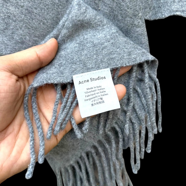 Op de afbeelding: Grijze sjaal met franjes. Het label leest "Acne Studios" en "Made in Italy" in meerdere talen. De sjaal is gemaakt van een zacht materiaal.