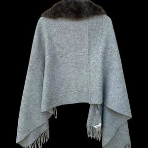 Op de afbeelding: Een grijze cape met een donkerbruine bontkraag hangt tegen een zwarte achtergrond. De cape heeft een gefranjerde zoom en is opgehangen aan een houten kleerhanger. De stof lijkt een zachte wolmix te zijn.