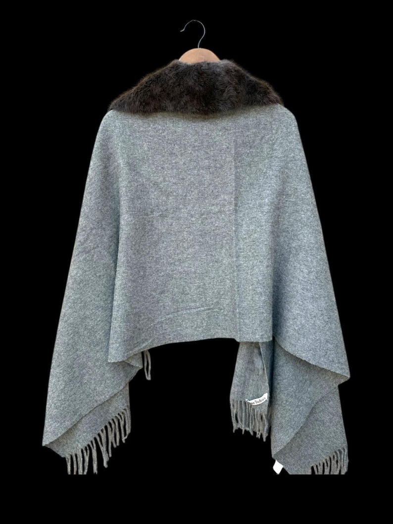 Op de afbeelding: Een grijze cape met een donkerbruine bontkraag hangt tegen een zwarte achtergrond. De cape heeft een gefranjerde zoom en is opgehangen aan een houten kleerhanger. De stof lijkt een zachte wolmix te zijn.
