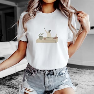 Puede incluir: Camiseta blanca con un gráfico de una persona sentada en una plataforma con una planta y un pequeño animal. La persona está extendiendo la mano hacia una mariposa.