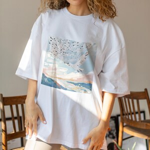 May include: White oversized t-shirt with a graphic print of a flock of birds flying over a landscape with a body of water and a sunset.