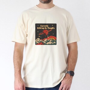 Könnte beinhalten: Ein cremefarbenes T-Shirt mit einer Retro-Grafik eines Planeten mit Ringen und Bergen. Der Text auf der Grafik lautet "Currently Orbiting My Thoughts".