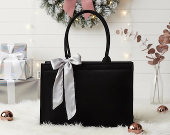 May include: A black tote bag with a silver ribbon bow. The bag has two handles and a rectangular shape. The background includes a Christmas wreath, wrapped gifts, and decorative baubles.