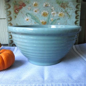 May include: A pale blue ceramic bowl with horizontal ridges. An orange pumpkin and a decorative gourd sit to the left of the bowl. The bowl is on a blue and white striped cloth, with a floral painting in the background.