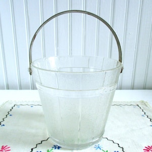 Puede incluir: Un cubo de hielo de vidrio transparente con un asa de metal plateado. El cubo tiene una superficie texturizada y un diseño acanalado.