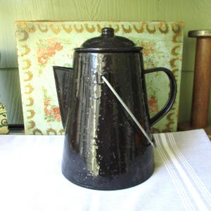 May include: A vintage black enamelware coffee pot with a lid, handle, and a white metal handle. The pot has a classic shape and is set against a backdrop of a floral-patterned box and a green wall. The pot is approximately 10 inches tall.