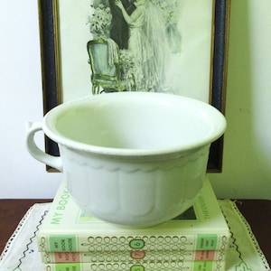 May include: White ceramic chamber pot with a single handle. The pot is sitting on top of three books with the text "My Book" visible on the top book.