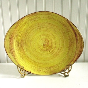 May include: A large, oval-shaped serving dish with a yellow glaze and a brown swirl pattern. The dish is on a gold metal stand.