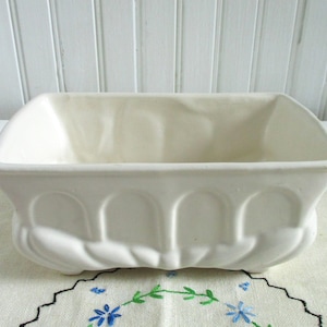 Peut inclure: Un pot de fleurs rectangulaire en céramique blanc cassé. Il présente un dessus incurvé et un motif en arc sur le devant, avec une base festonnée. Le pot vide repose sur un tissu brodé de fleurs. Dimensions en centimètres.