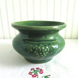 May include: A green ceramic planter with a grape design. The planter is glazed and has a rounded shape.