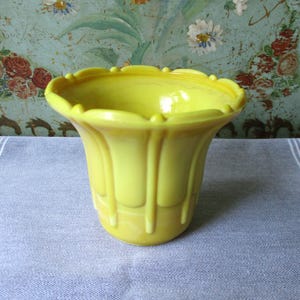 May include: A bright yellow, vintage-style vase with a fluted design and scalloped rim. The vase is made of a glossy material and sits on a gray and white striped surface. The background features a floral pattern.