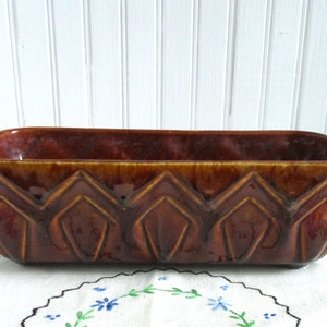May include: A brown glazed ceramic planter with a rectangular shape and a geometric design. The planter has a slightly raised rim and a textured surface.